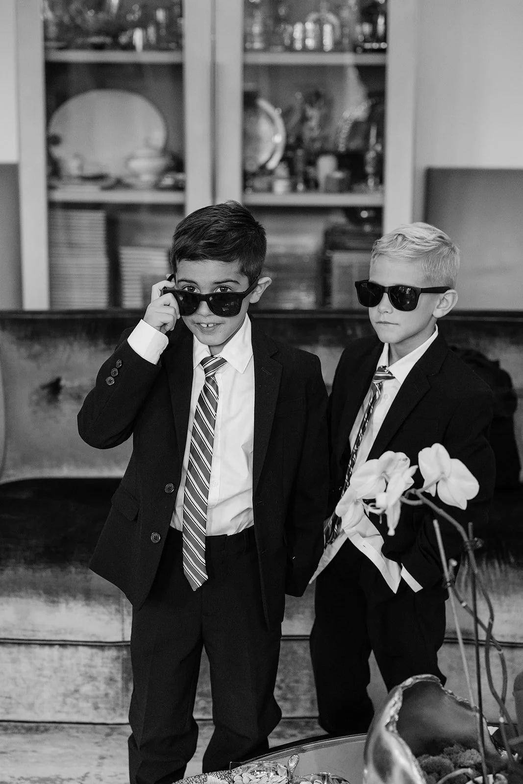 Black and white photo of two boys in suits and sunglasses, one holding white calla lilies