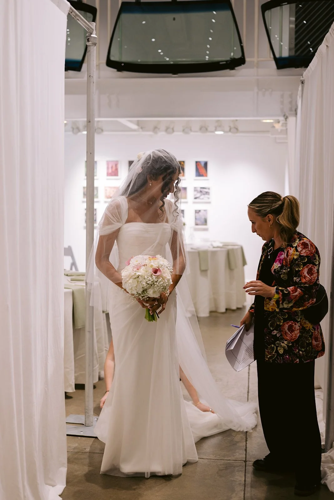 A bride in an ivory gown holds her bouquet as a woman adjusts her veil behind white drapery