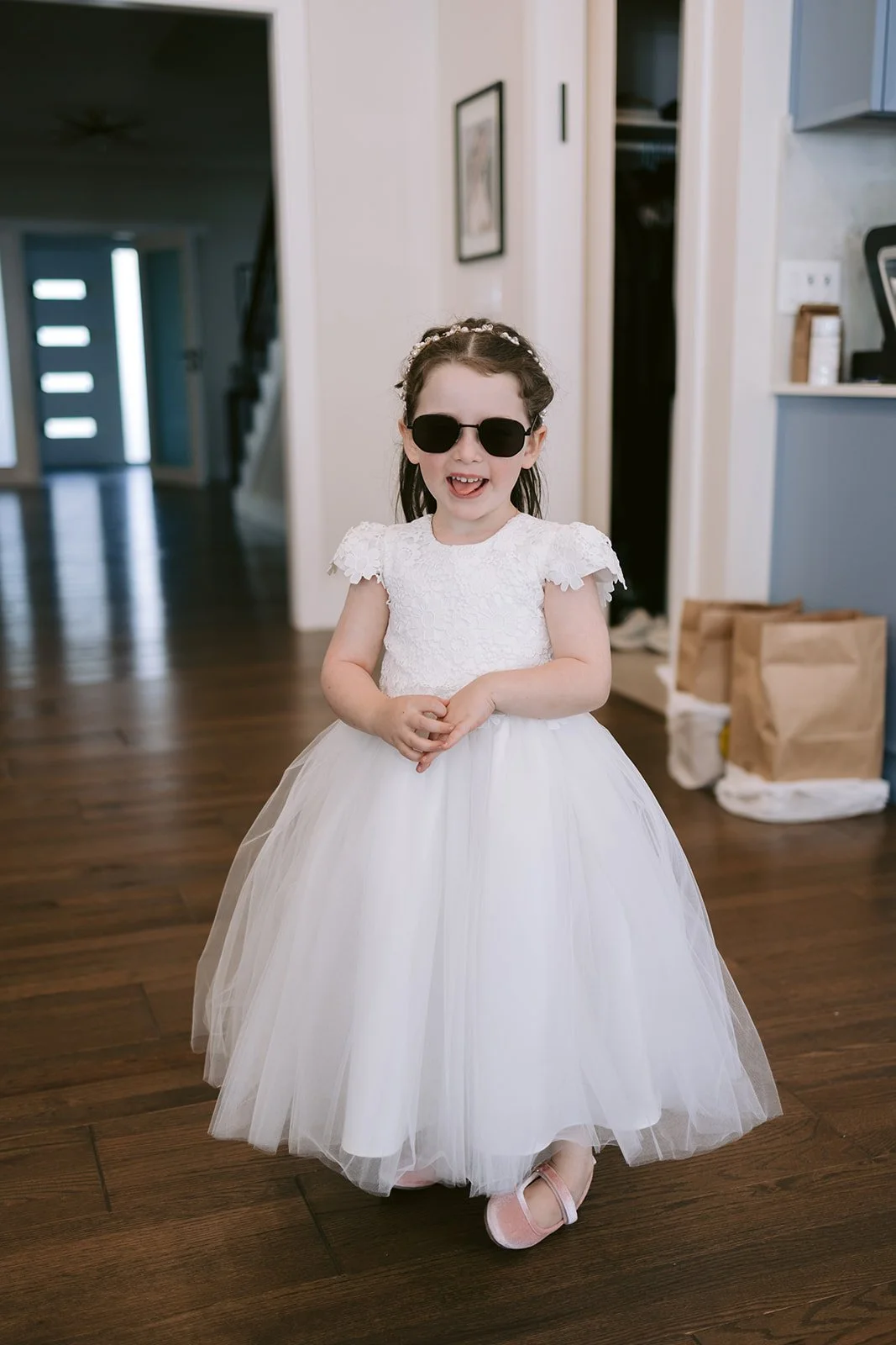 A flower girl in a white tulle dress and sunglasses smiles in a hallway