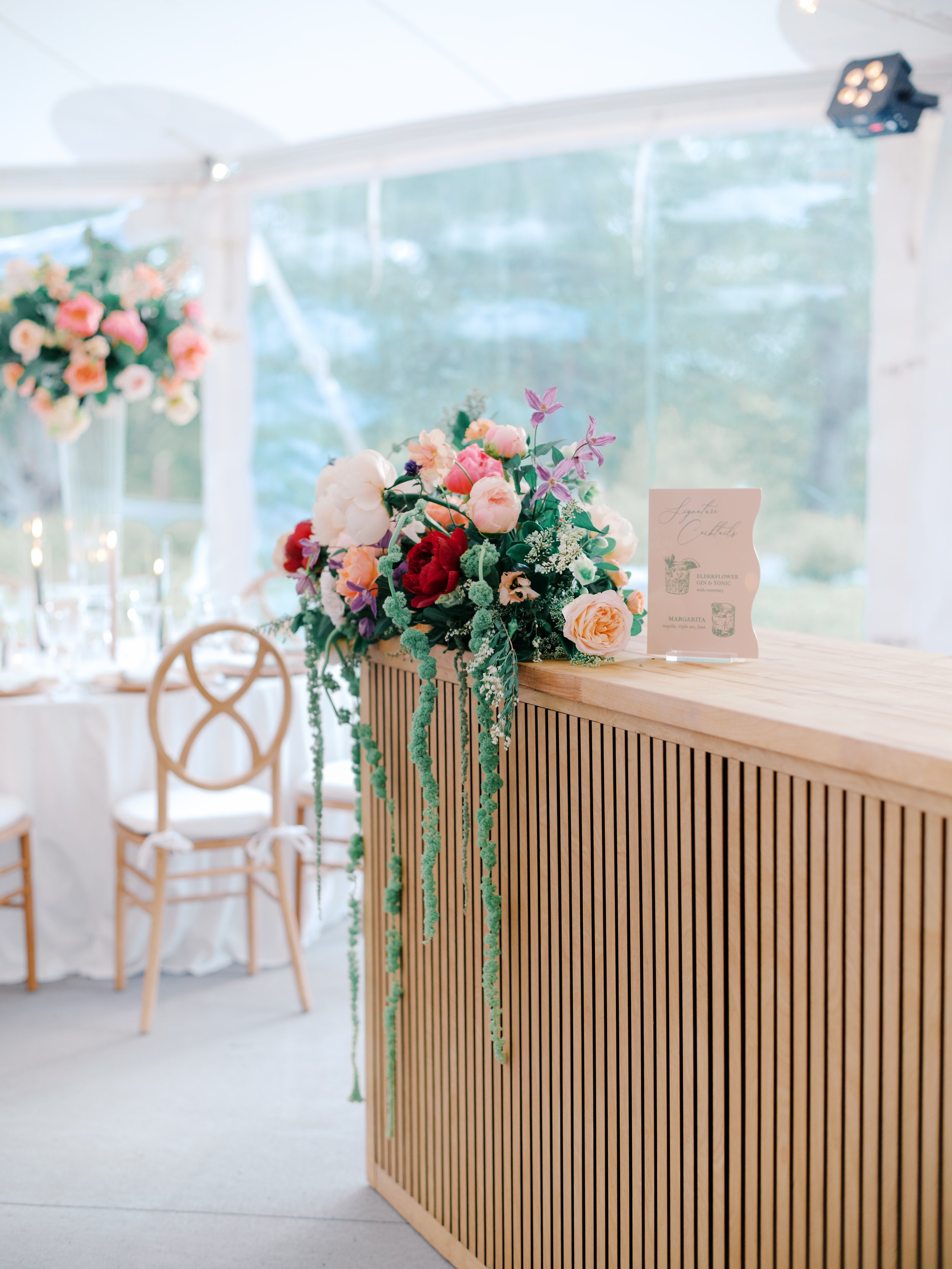 A slatted wood bar topped with a cascading floral arrangement inside the wedding tent