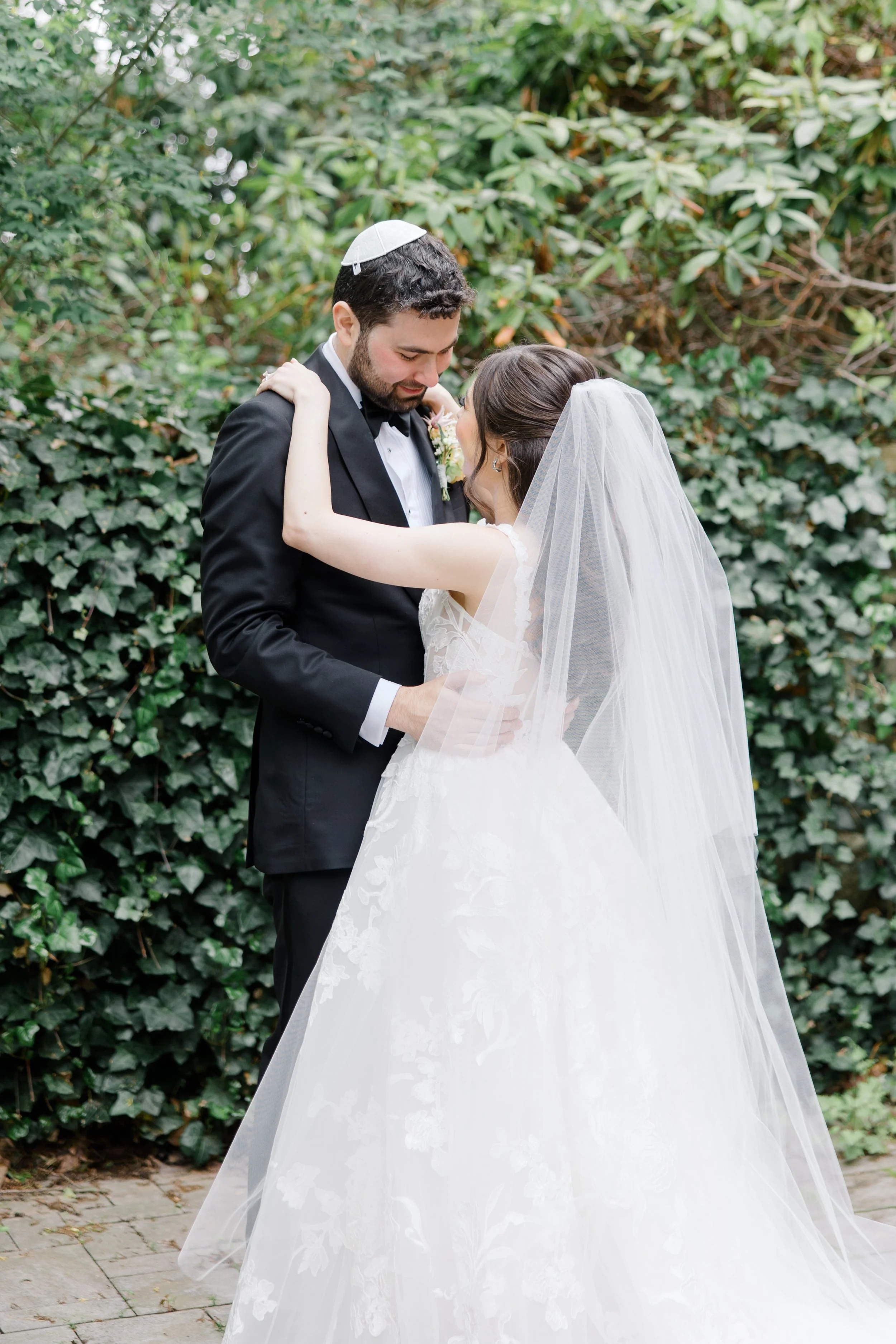 The couple embraces in front of an ivy-covered wall