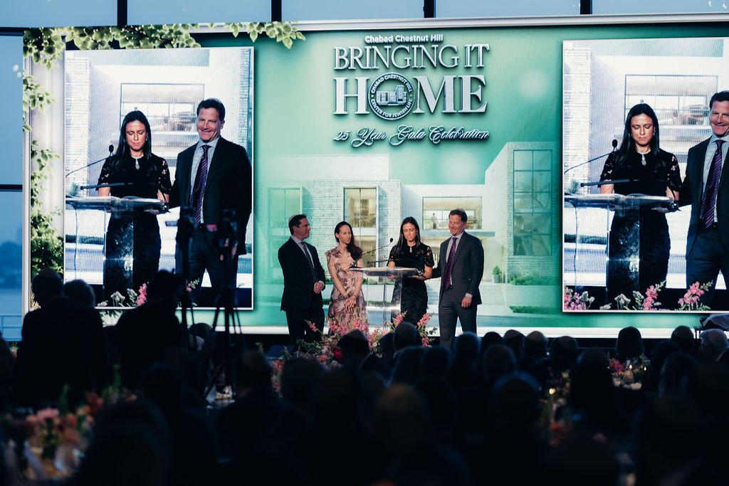 Presenters on stage beneath a Bringing It Home screen at the Chabad Chestnut Hill 25th anniversary gala
