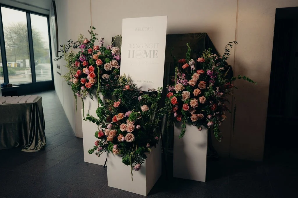 Floral arrangements on white pedestals flank the Bringing It Home sign beside the windows