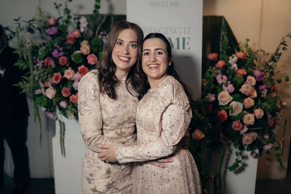 Two women in ivory dresses embrace beside tall floral arrangements at the gala entrance