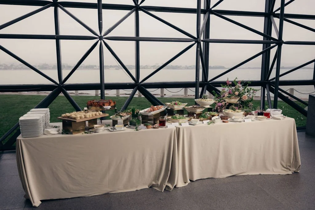A buffet table set before the atrium's steel-framed windows overlooking the water