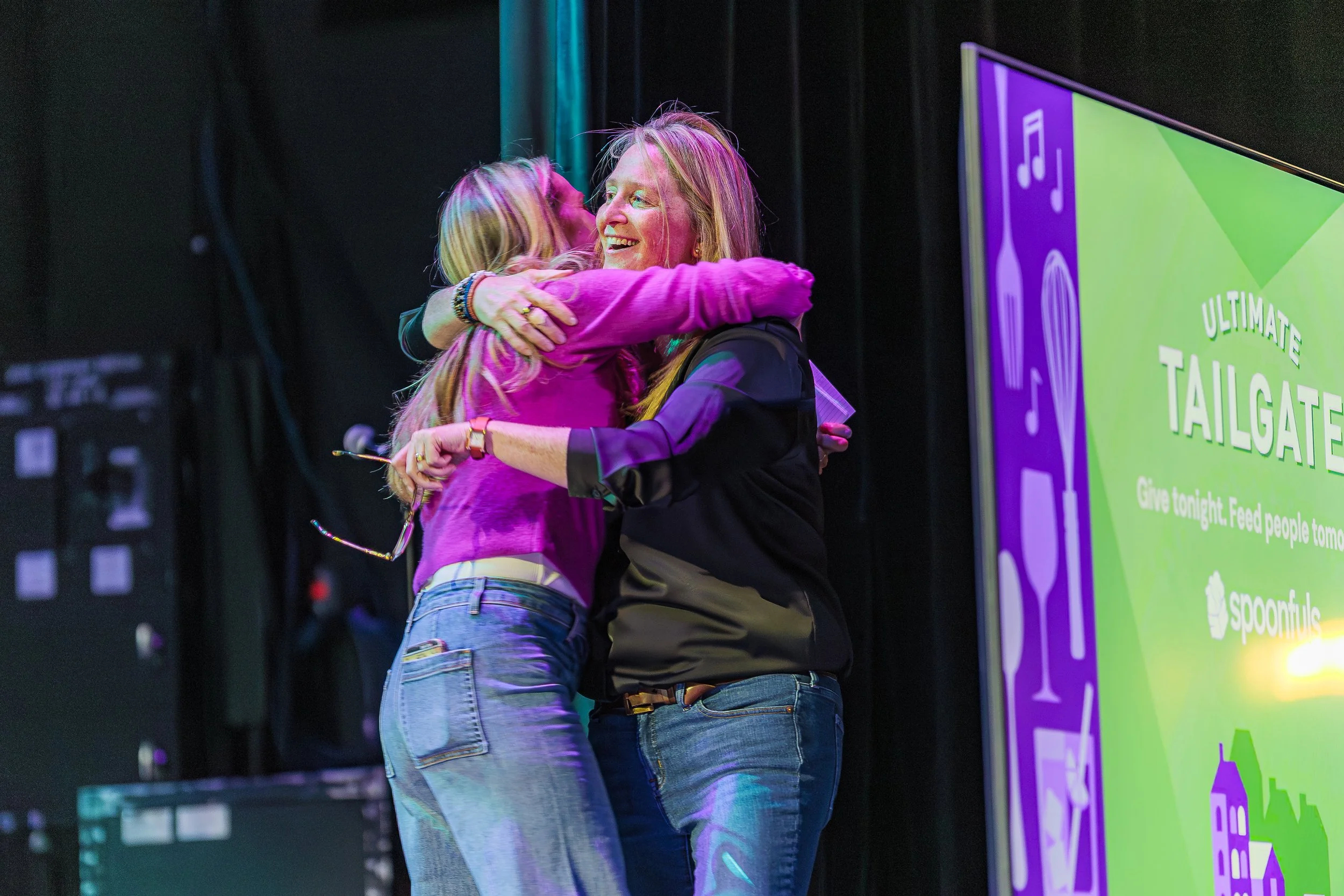 Two women embrace on stage beside the Ultimate Tailgate screen at the Spoonfuls gala