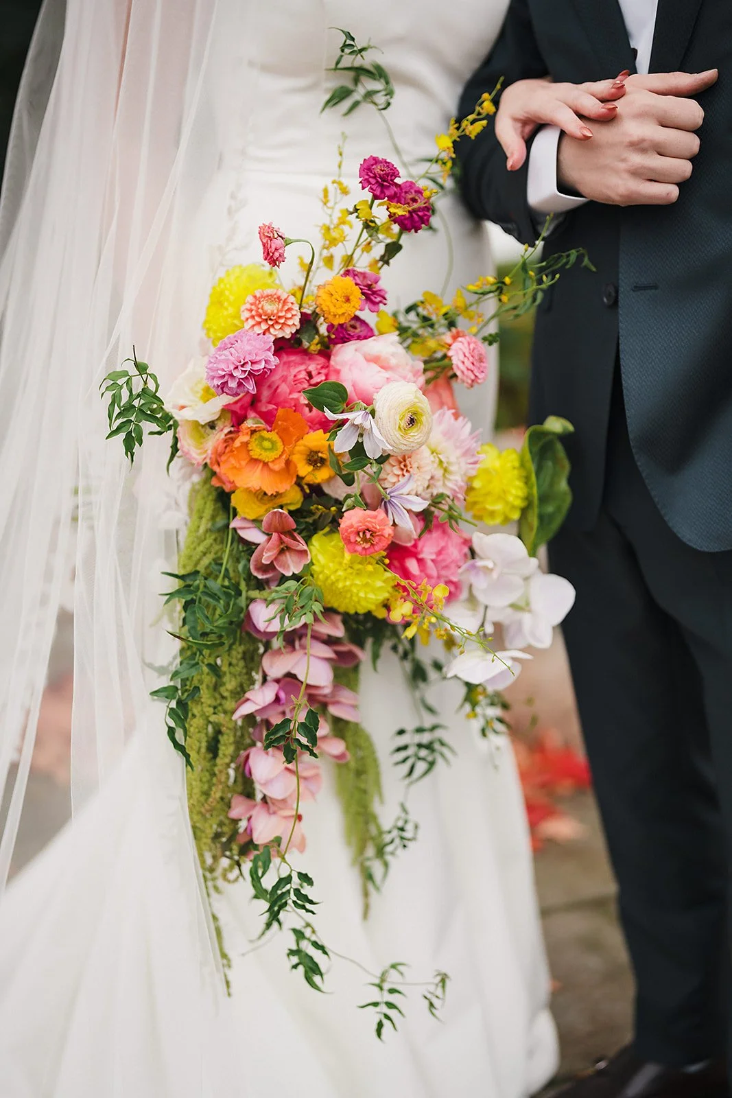 The bride holds a cascading bouquet of bright pink, orange, and yellow blooms beside the groom
