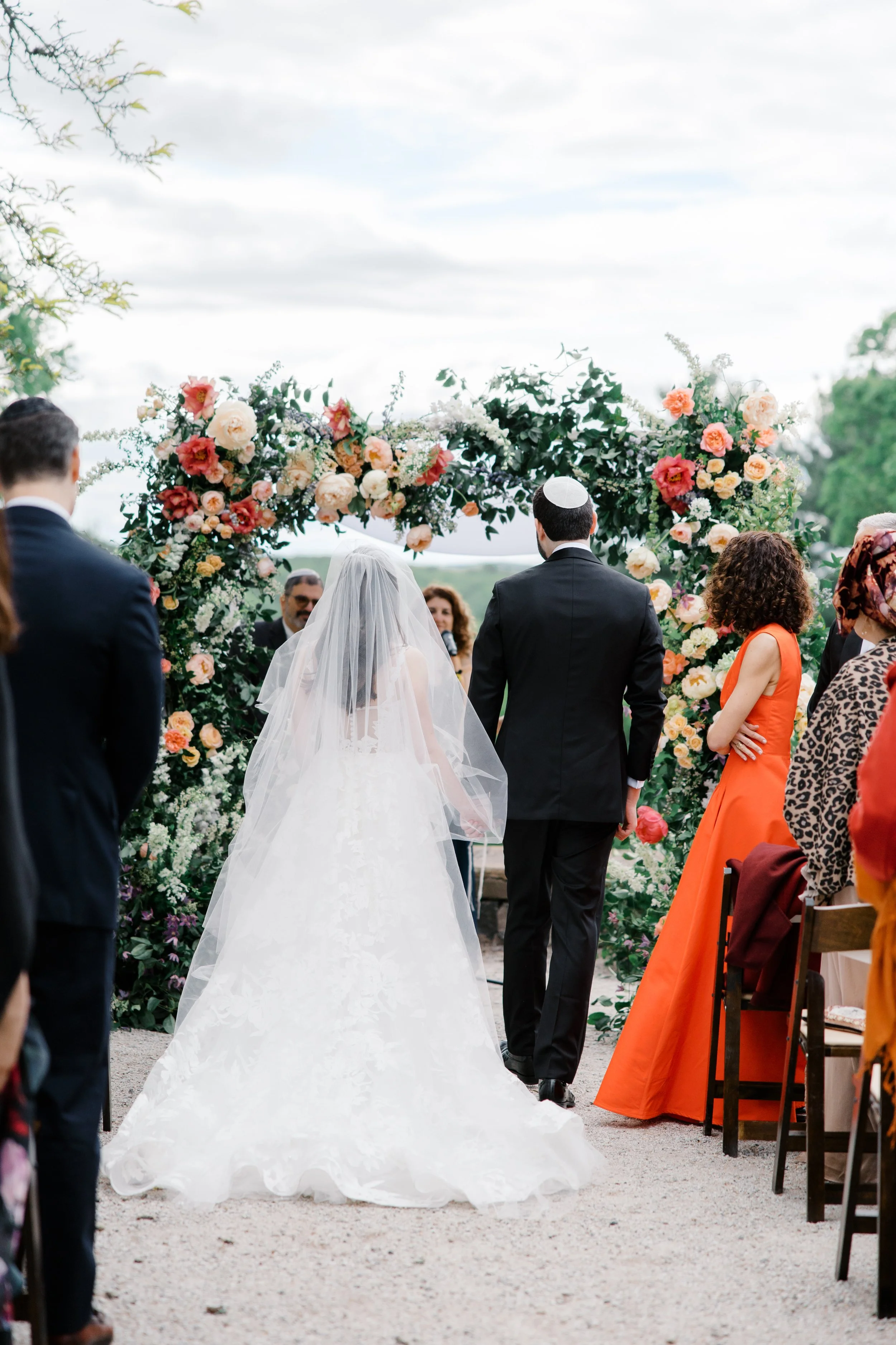 The bride and groom stand under a floral chuppah, seen from behind during the ceremony