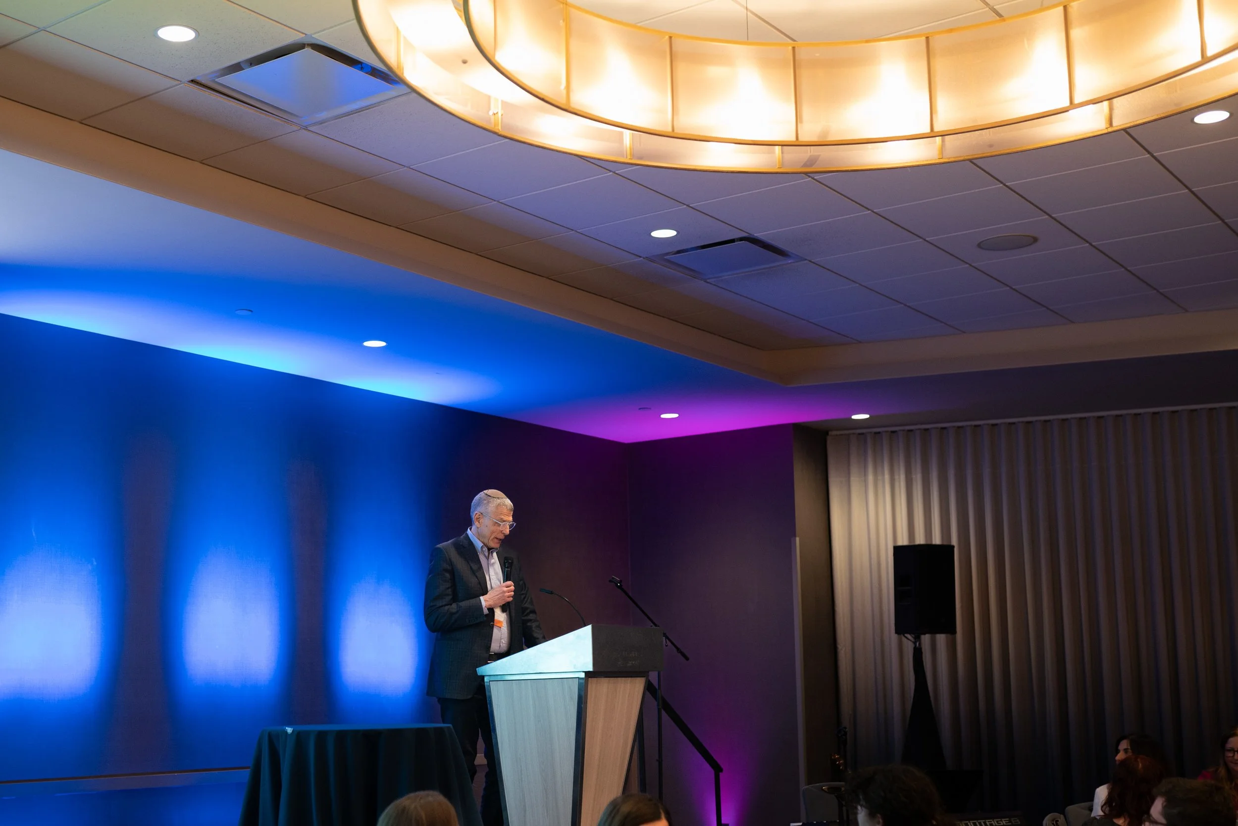 A speaker at a podium addresses the ballroom under blue stage lighting at the ARJE conference