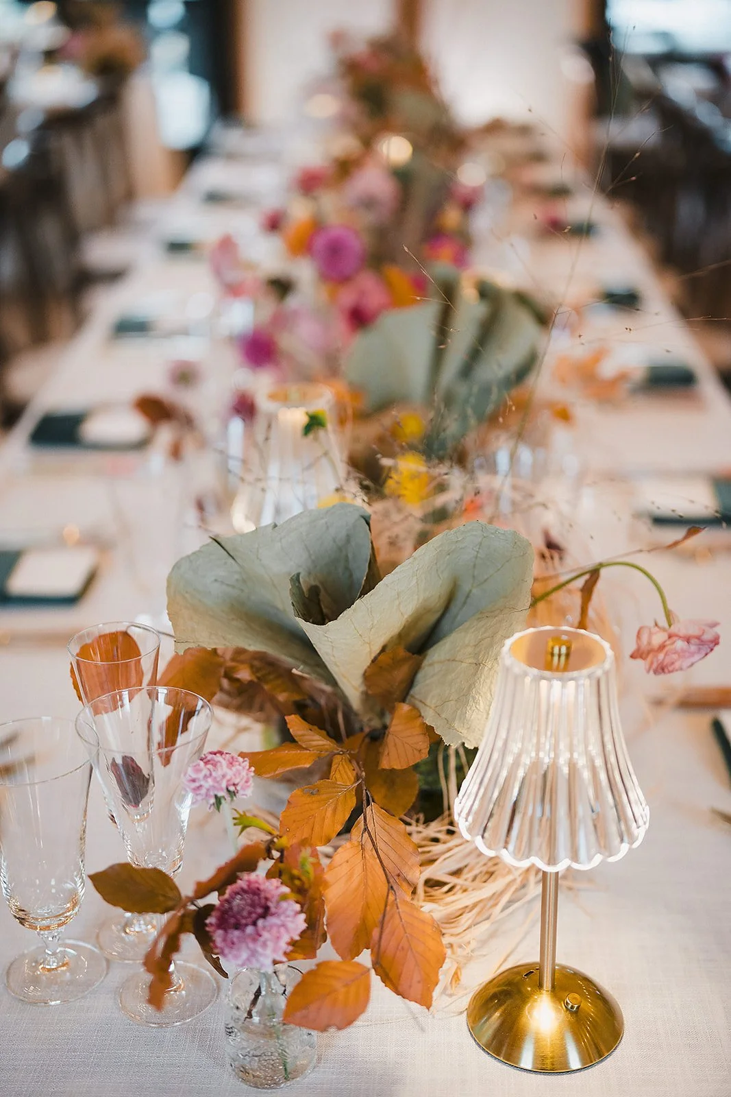A tablescape of autumn leaves, dried florals, and a brass table lamp running down a long table