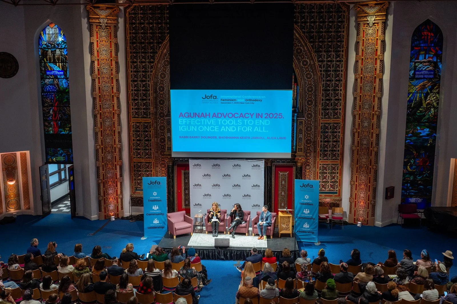 Wide view from the balcony of a packed sanctuary as a panel speaks beneath a large screen at the JOFA conference