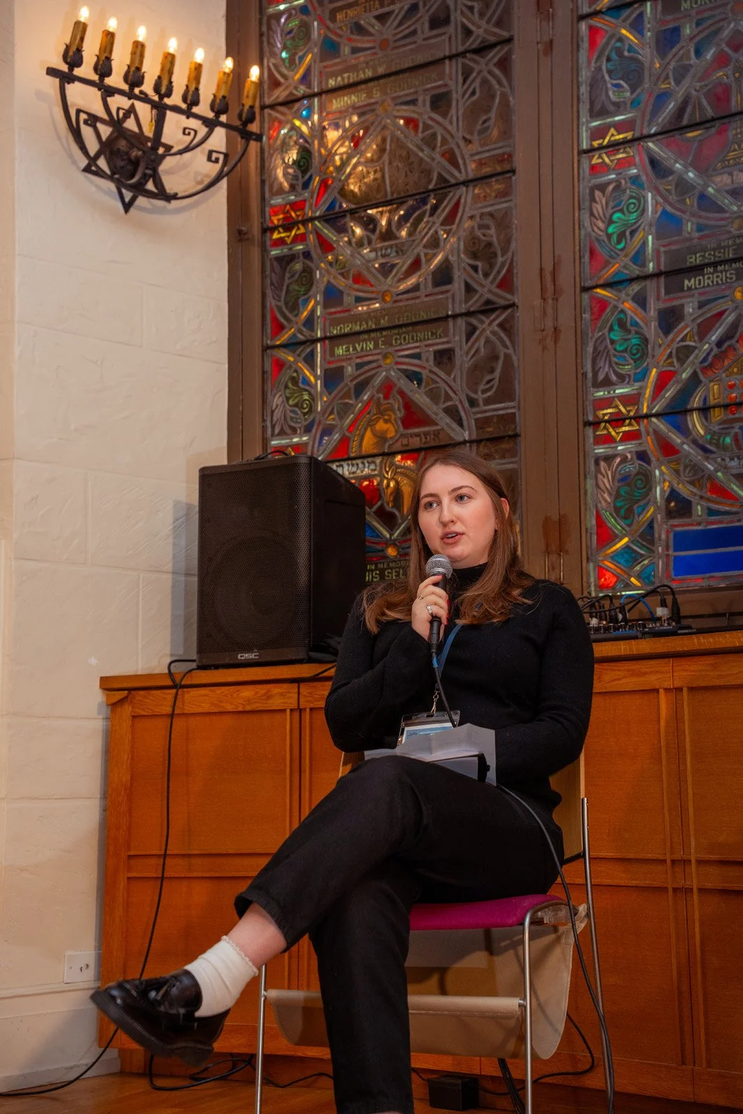 A speaker with a microphone addresses a session in front of stained glass at the JOFA conference