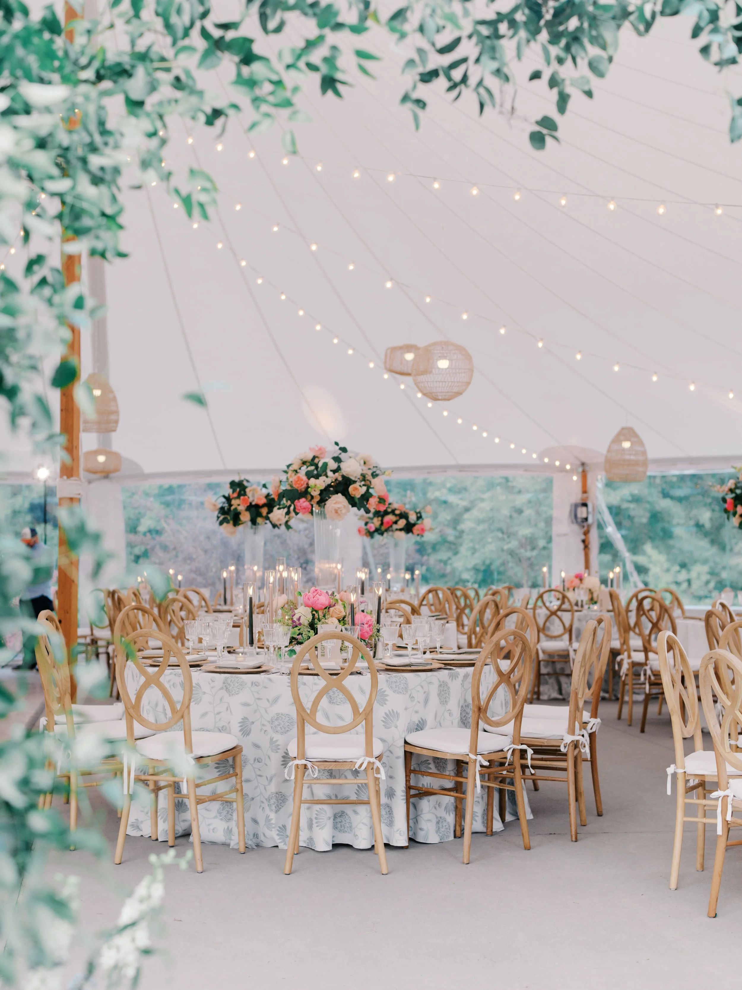 Round tables with gold chairs and tall floral centerpieces under string lights in the tent