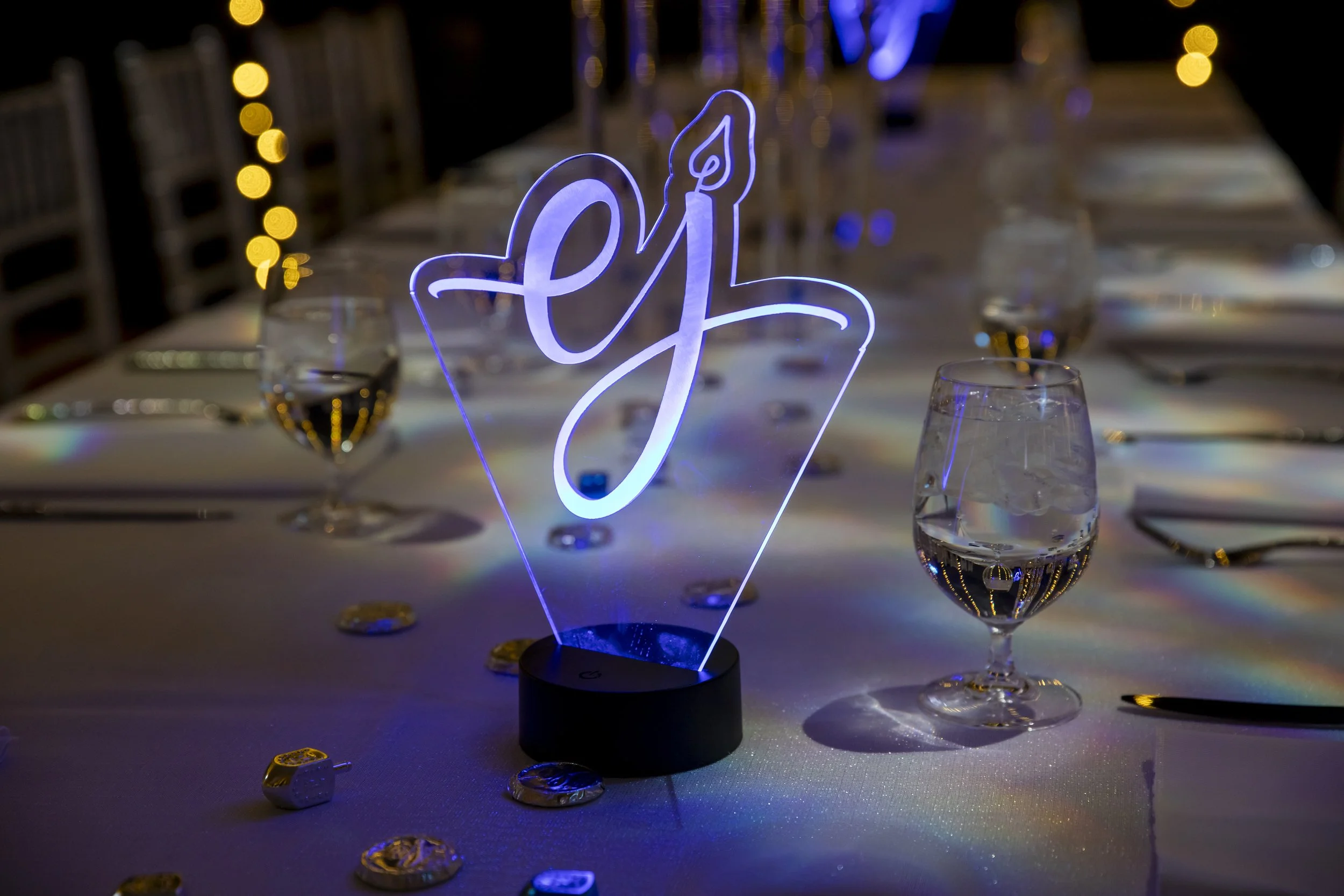 A glowing acrylic EJ monogram with a candle flame lights up a dinner table at Eloise's bat mitzvah