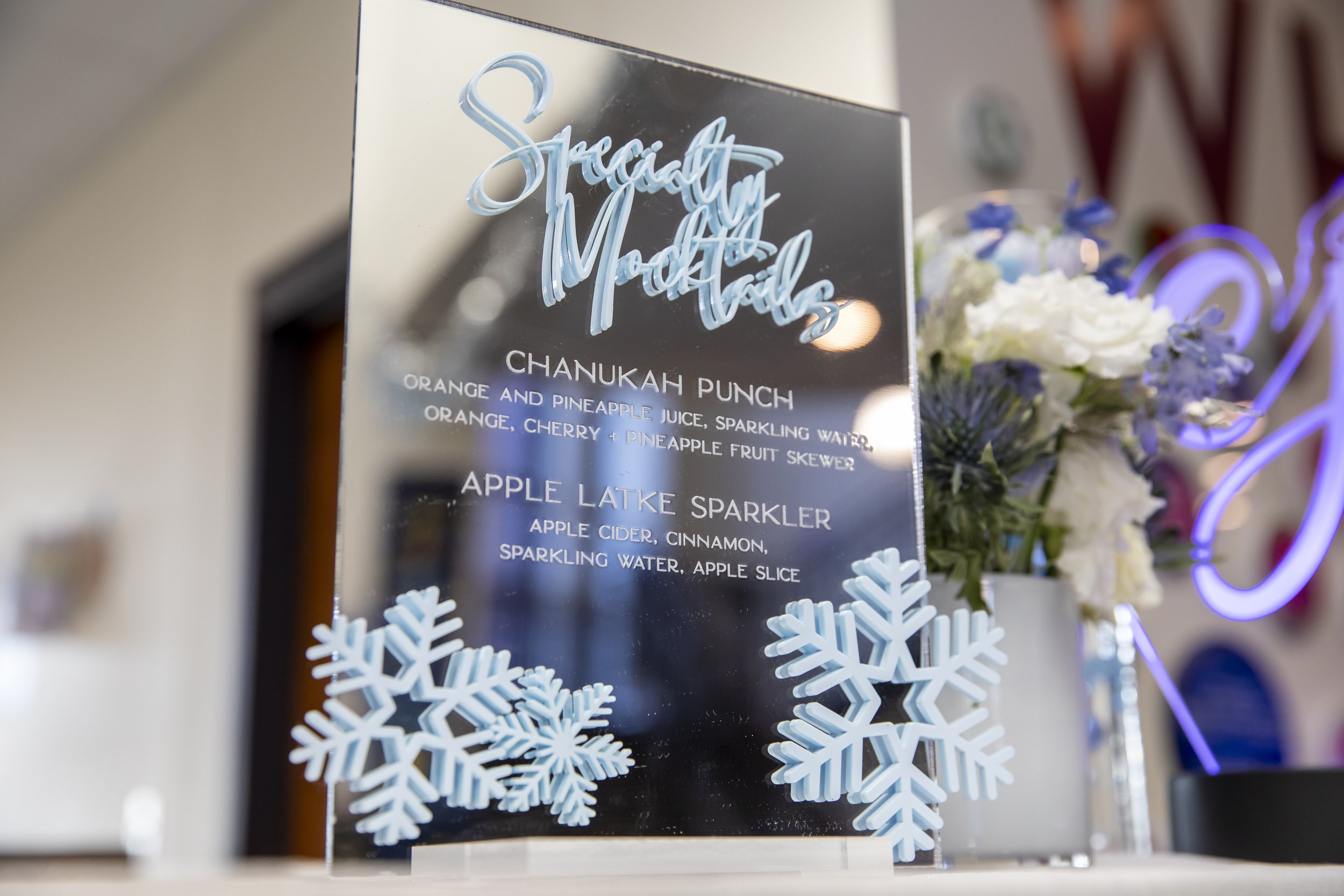 An acrylic specialty mocktails sign listing a Chanukah punch and an apple latke sparkler, trimmed with blue snowflakes