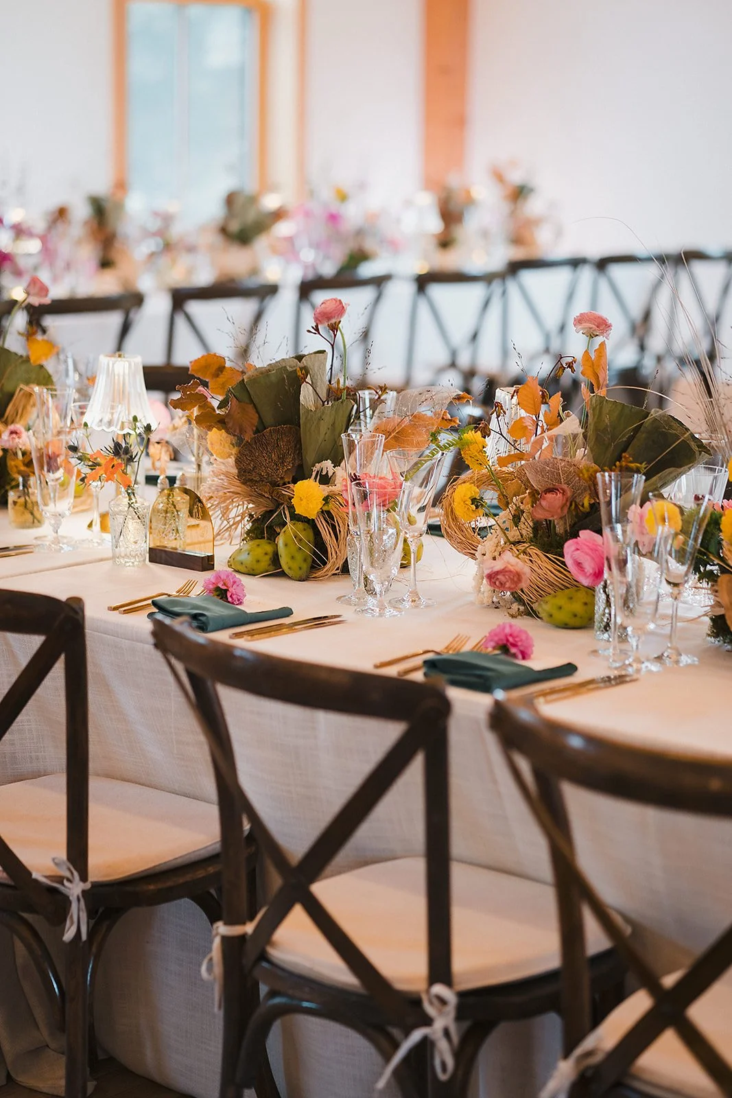 Long tables with dark cross-back chairs and colorful floral runners in a sunlit barn