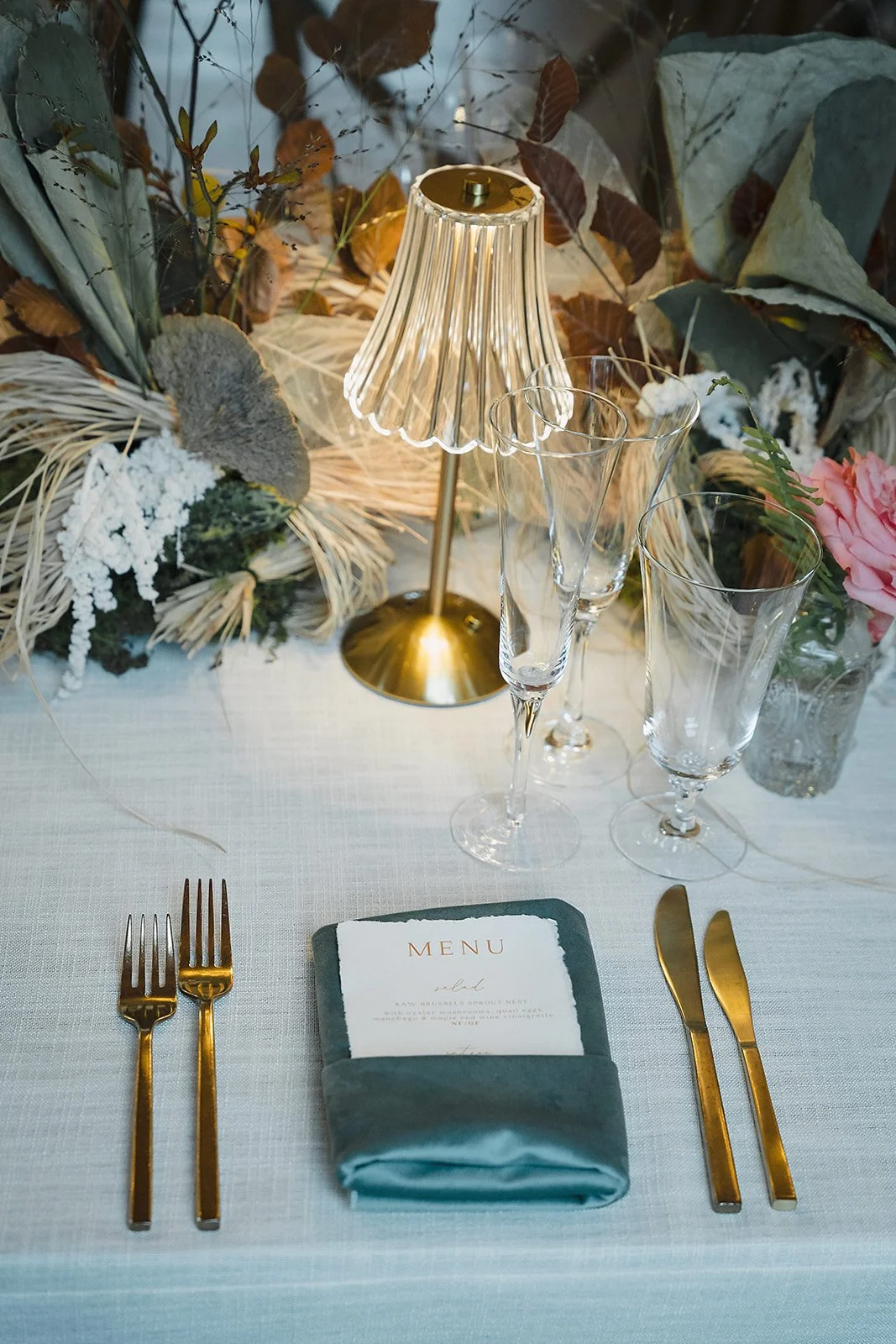 A place setting with a dusty blue napkin, gold flatware, menu card, and a glowing glass lamp