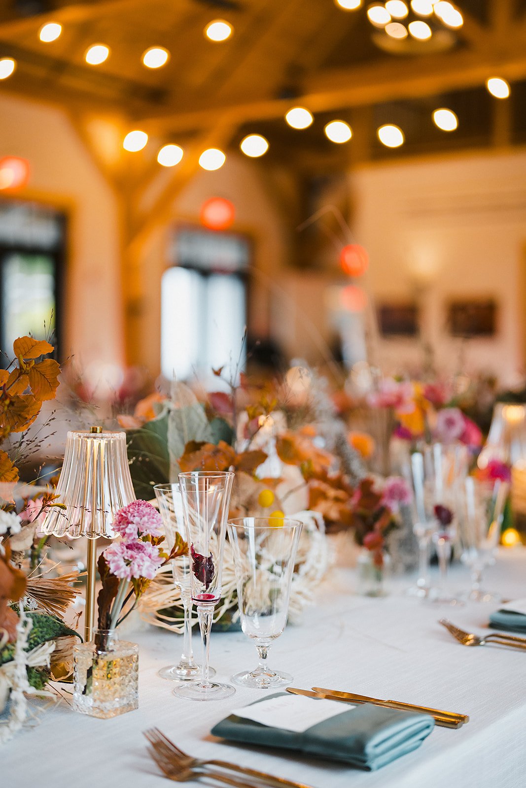 A long table set with dried and fresh florals, glassware, and green napkins beneath string lights in a barn