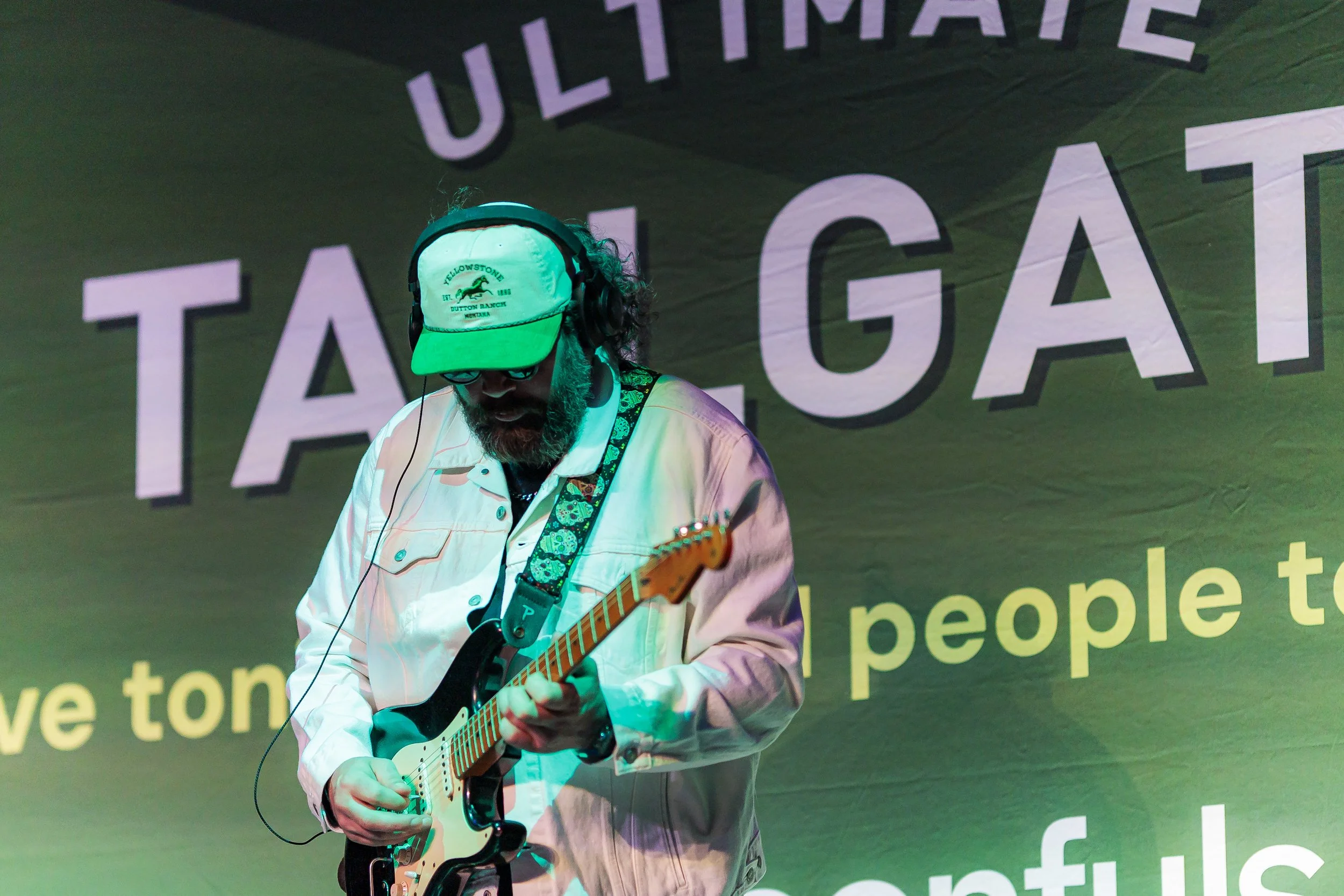 A guitarist in a green cap performs in front of the glowing Ultimate Tailgate backdrop