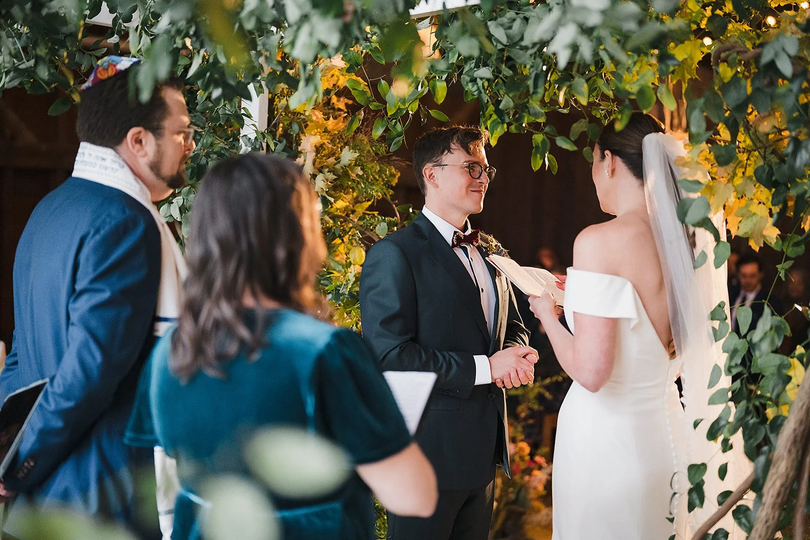 The groom smiles as the couple exchanges vows beneath a greenery-covered arch