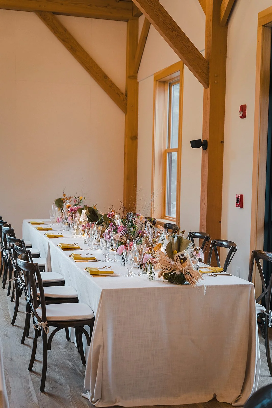 A long reception table with a champagne linen and dark cross-back chairs in a timber-framed room