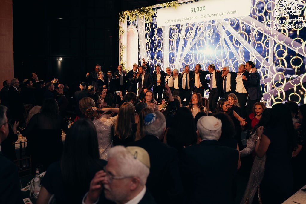 Guests applaud as a group stands on a stage lit by a sparkling geometric backdrop during the fundraising appeal