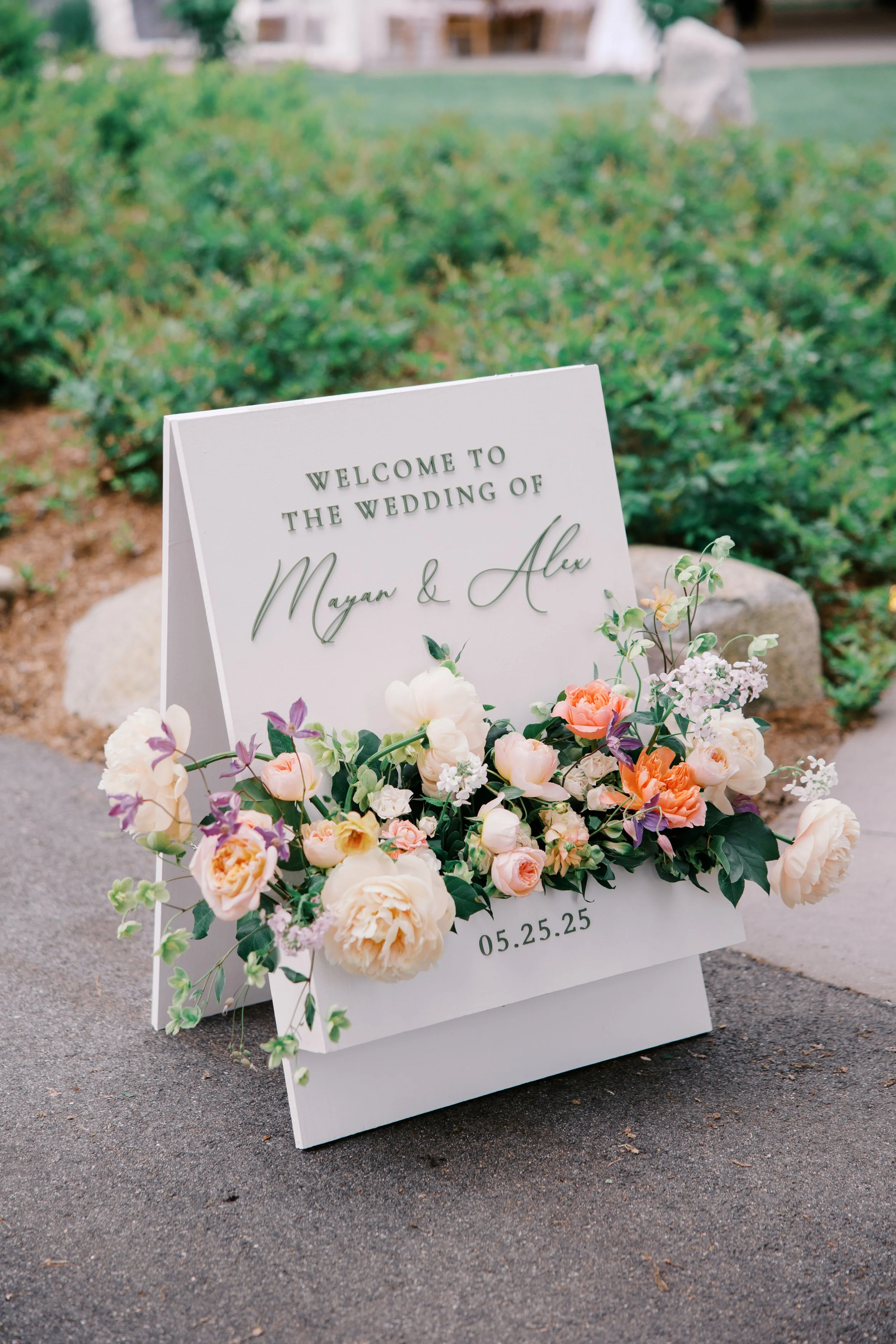 A welcome sign for the wedding of Mayan and Alex trimmed with peach and pink flowers
