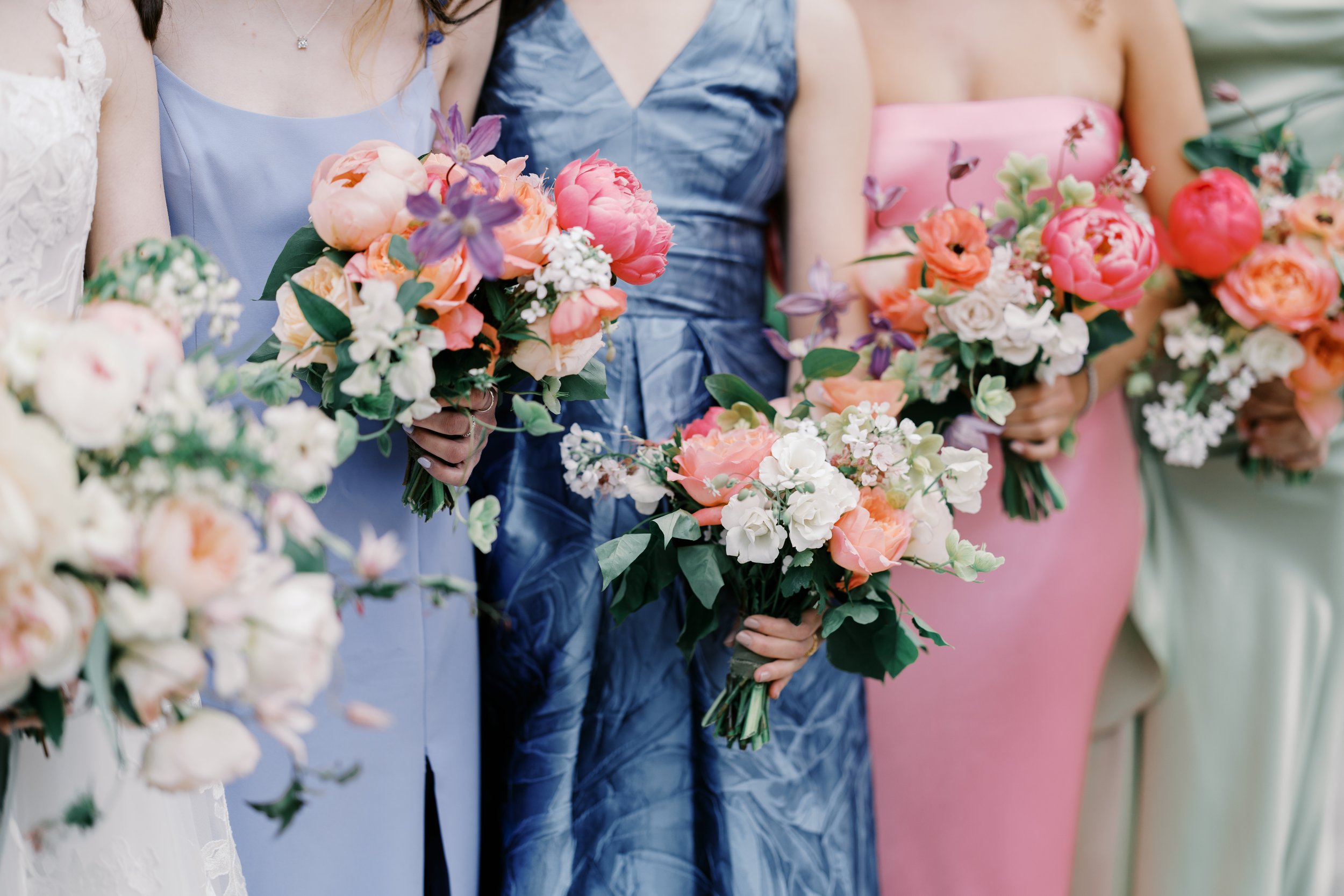 Bridesmaids in mismatched pastel dresses hold bouquets of pink and peach blooms