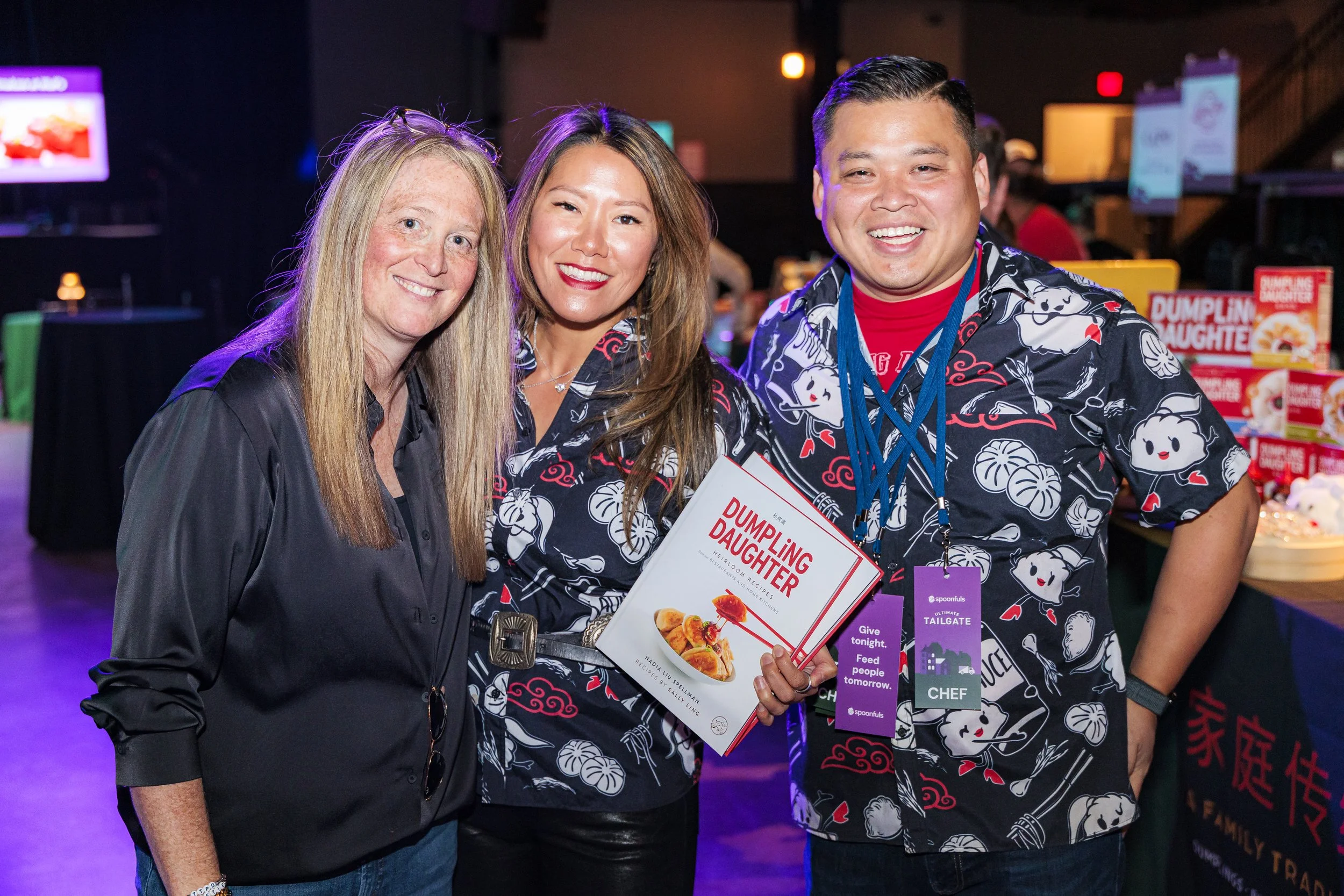 Three guests pose together, one holding a cookbook, at the Spoonfuls Ultimate Tailgate