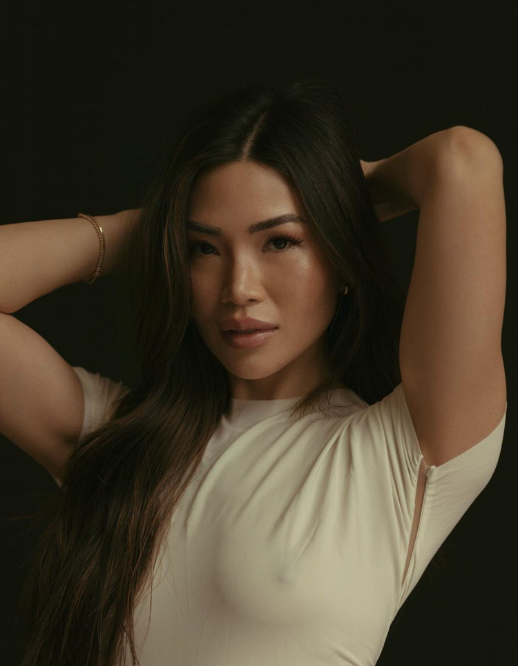 A woman with long dark hair posing with her arms behind her head, wearing a cream-colored top against a dark background.