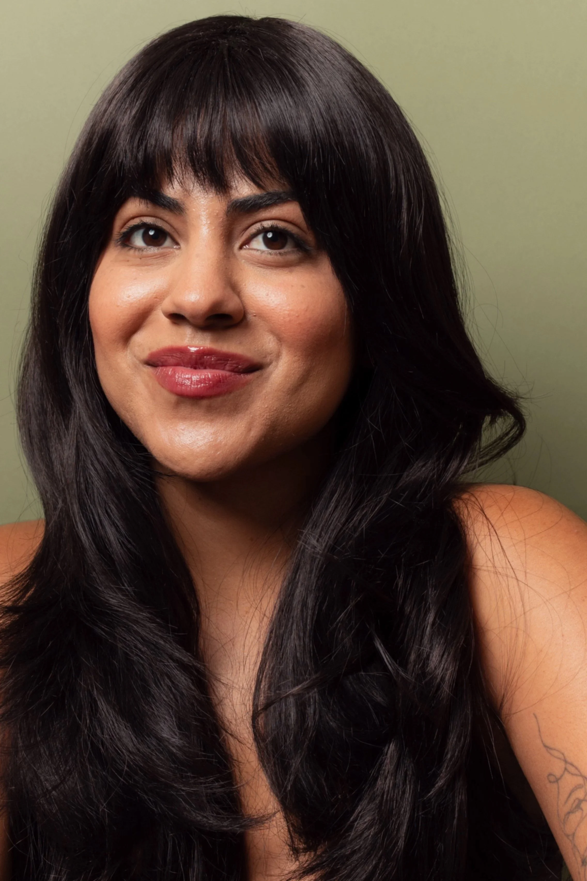Close-up of a woman with long, dark hair and bangs, smiling slightly, against a plain, light green wall.