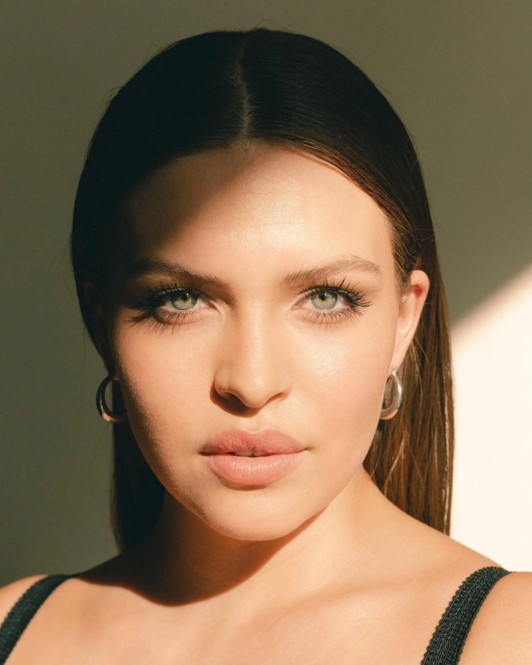 Close-up of a woman with straight brown hair, green eyes, wearing a black strap top, and earrings.