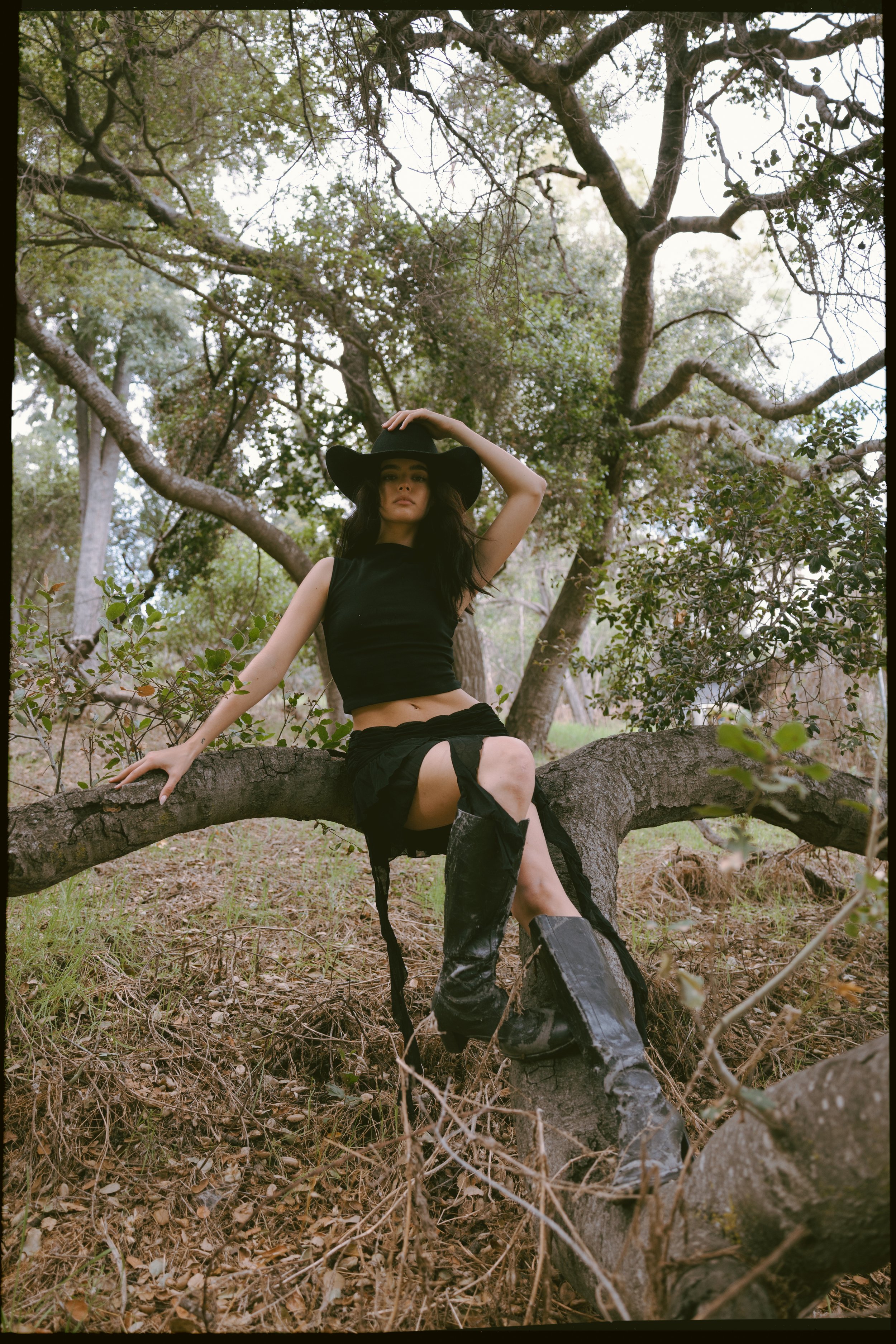 A young woman dressed in black, wearing a wide-brimmed hat, sitting on a large fallen tree in a forested area with many trees and green foliage around her.
