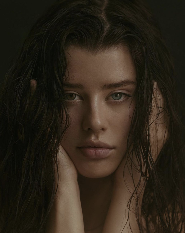 A close-up portrait of a woman with wet, dark hair, resting her face softly on her hands, looking directly into the camera against a dark background.
