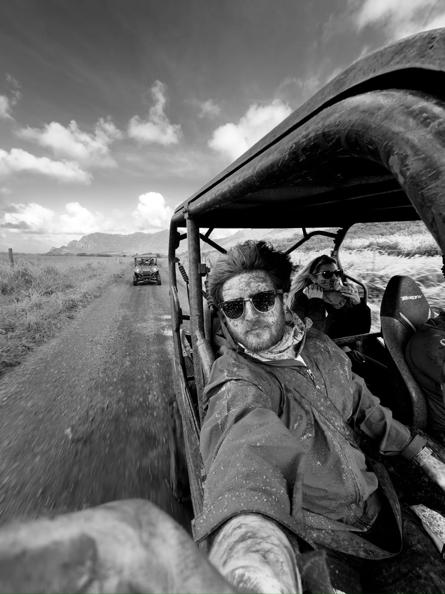 Black-and-white photo of three people riding in an off-road vehicle on a dirt road in a rural landscape with mountains in the background and a cloudy sky.