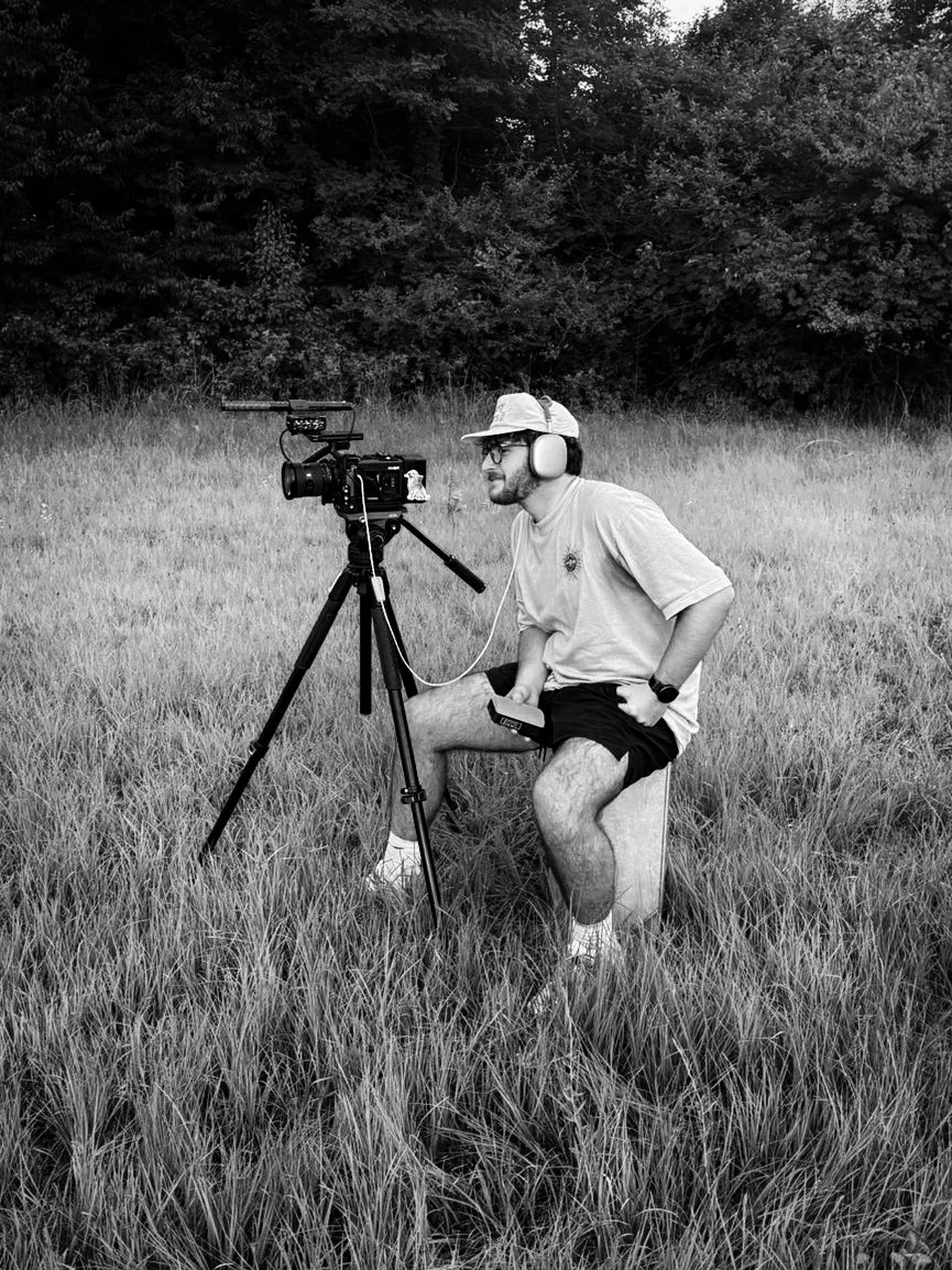 A man filming outdoors in a grassy field with a professional video camera on a tripod, wearing headphones, a hat, glasses, and casual clothing.