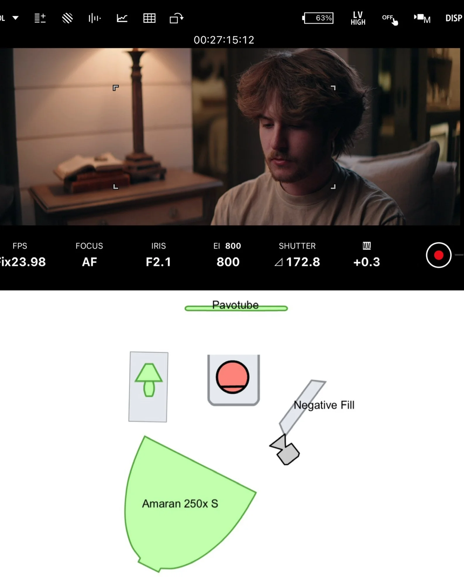 The image shows a camera screen capturing a scene with a young man with voluminous, wavy brown hair, sitting indoors next to a lit table lamp and books. Overlaid on the camera interface are various controls and settings such as battery life, focus, shutter speed, ISO, and a recording indicator.