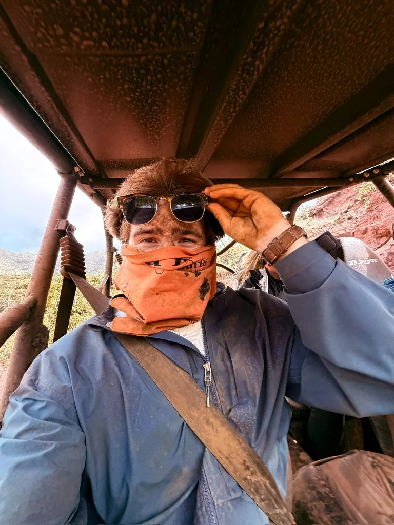 A man wearing a blue jacket, dust on his clothing, a brown strap across his chest, and a brown face covering. He is holding sunglasses with his right hand, under an open-air metal structure with a mountain landscape in the background.