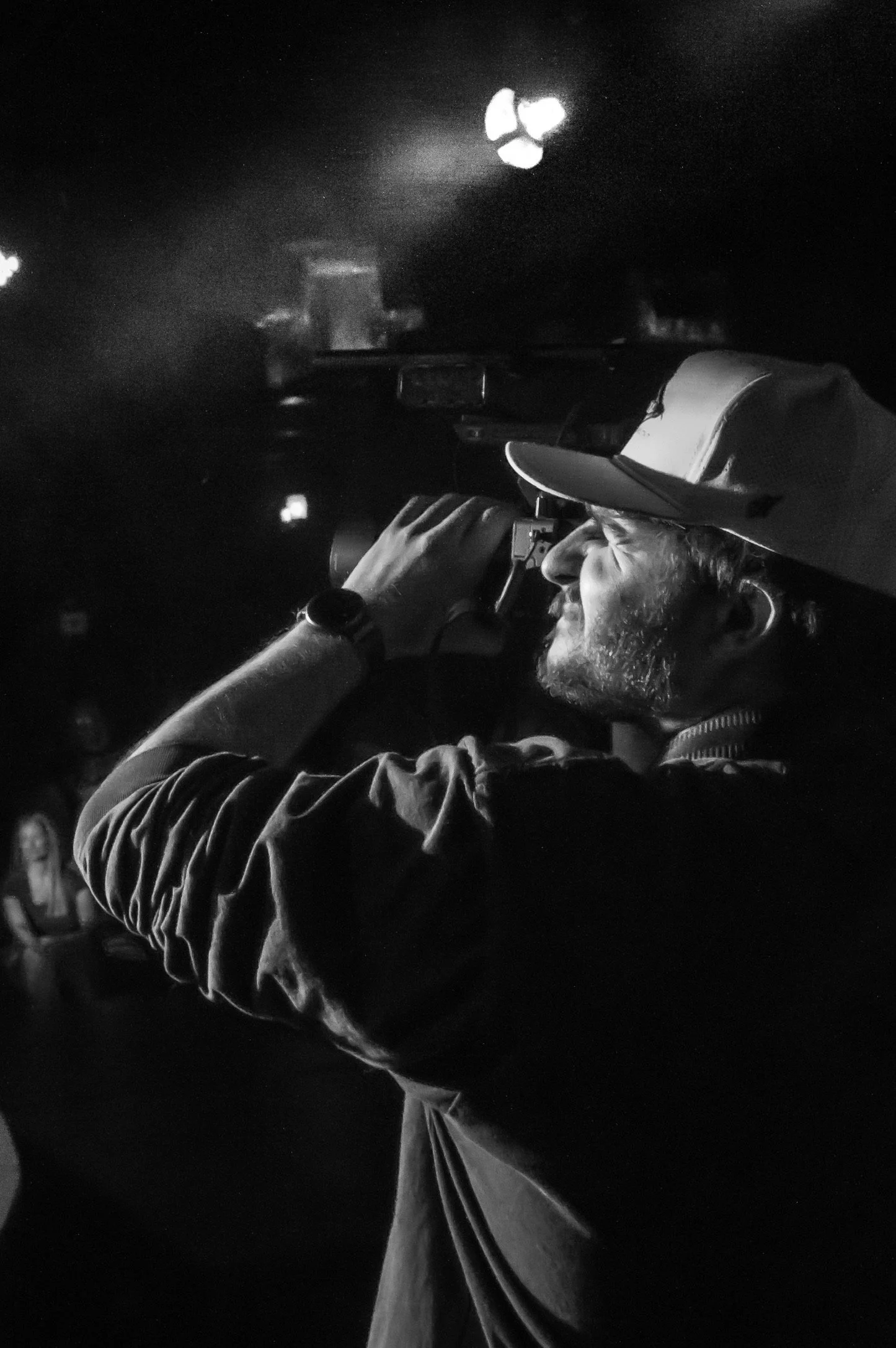 A man wearing a cap and a watch is looking through binoculars at night. The background is dark with some faint lighting.