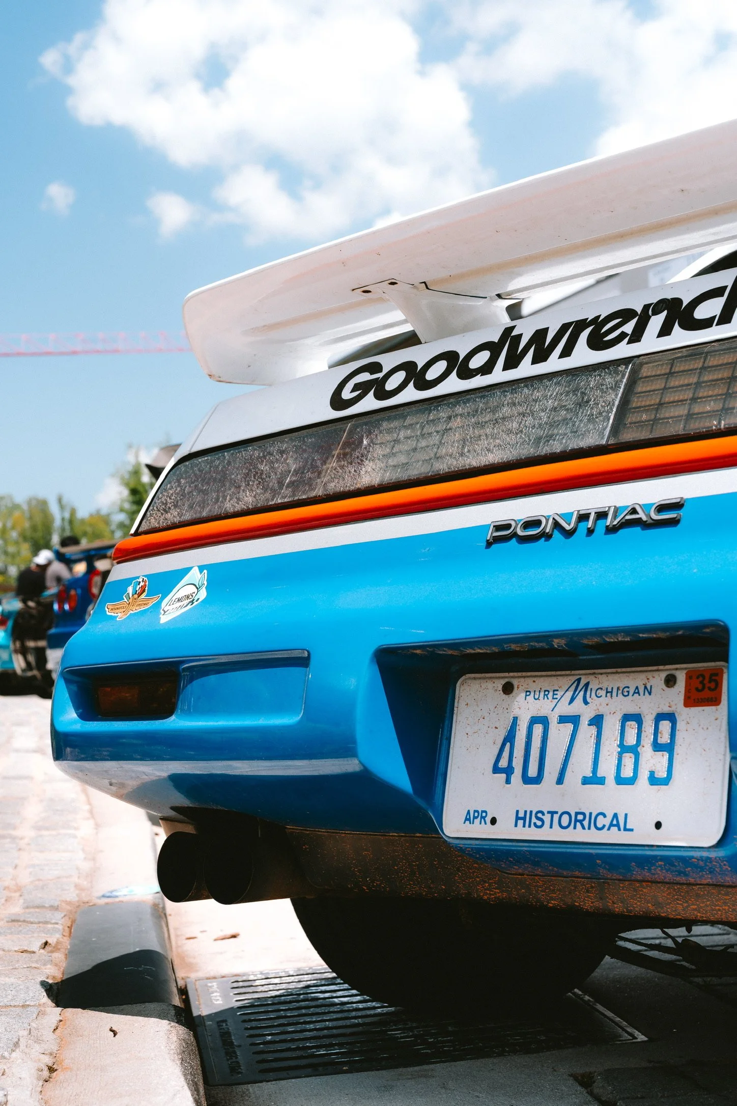 Rear view of a blue Pontiac race car with a white rear wing, parked on a city street with a drain grate and a curb, under a partly cloudy sky.