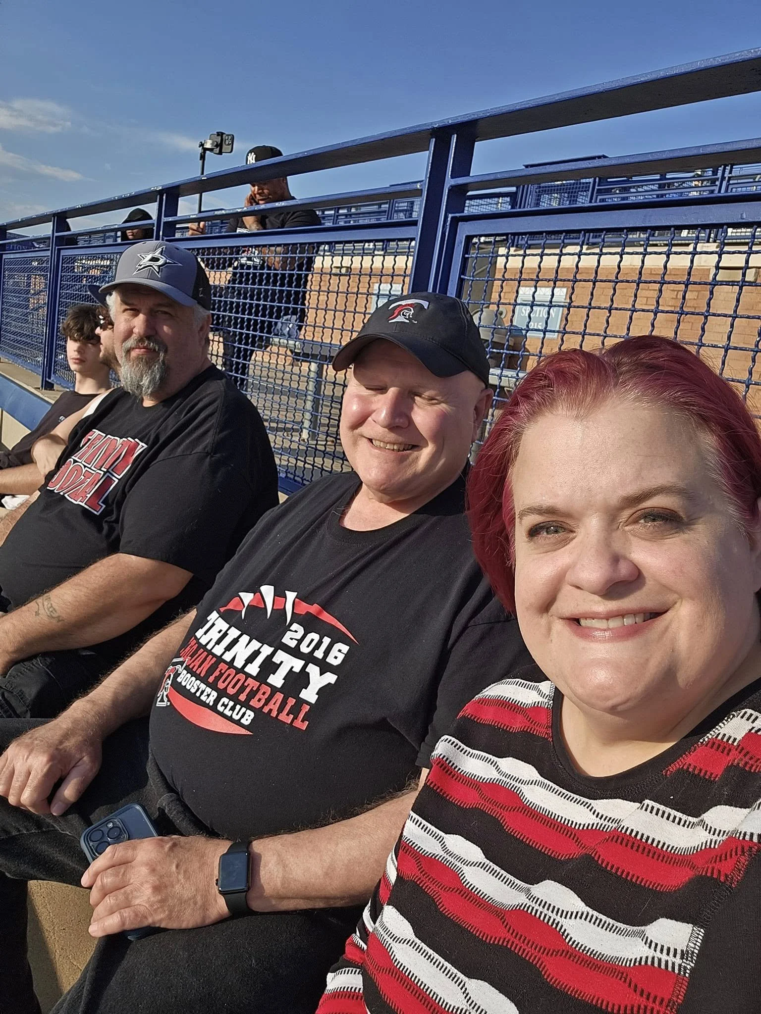 Group of four people sitting on bleachers at a high school football game, with a blue sky overhead. Three of the individuals are wearing black t-shirts, and one woman has red hair and is wearing a red and white striped shirt.