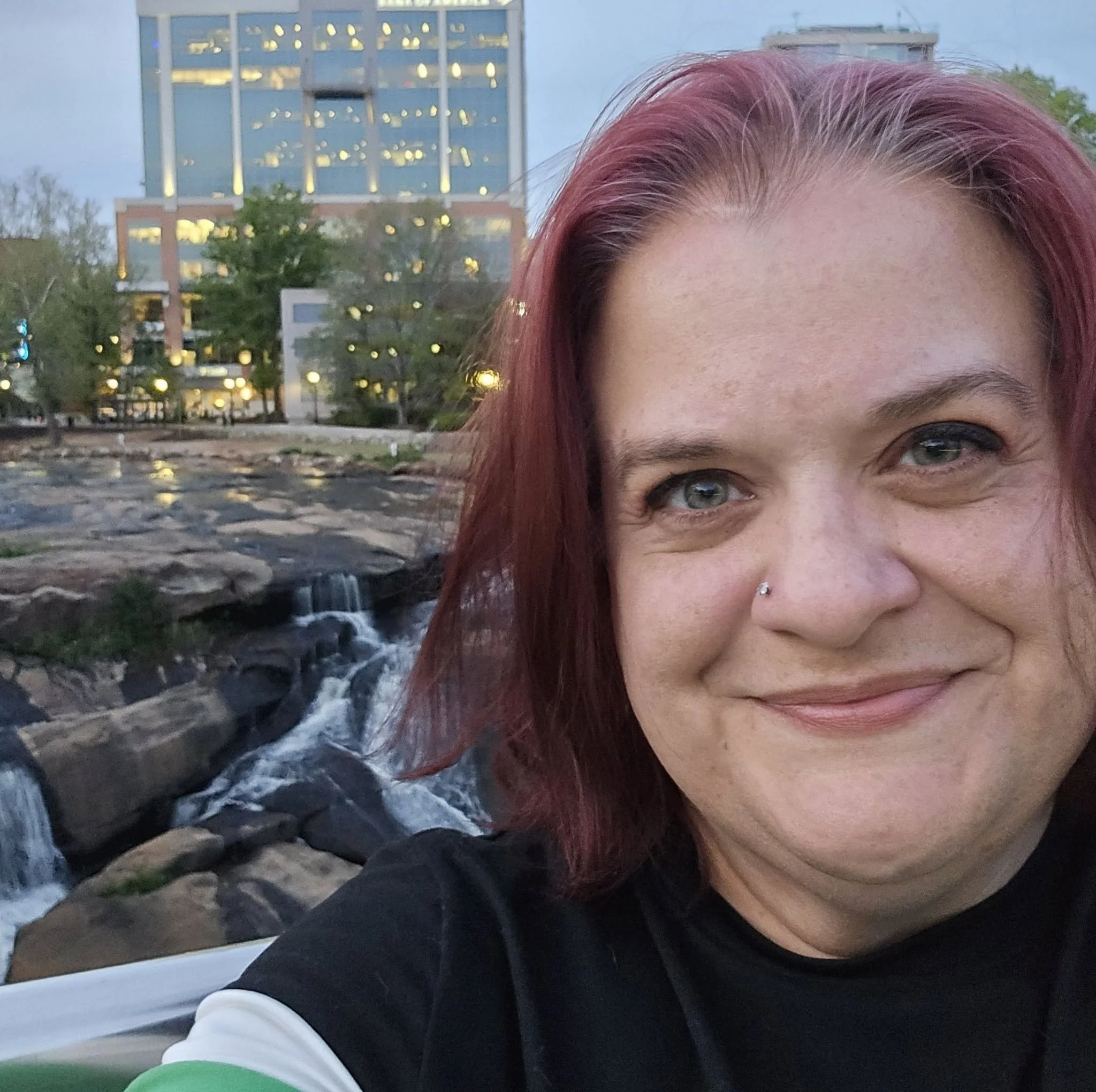 Close-up of a woman's face and eye, with background of a modern glass building, trees, and a small waterfall with rocks.