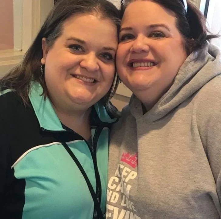 Two women smiling for a selfie, close together indoors, one wearing a black and teal jacket and the other in a gray hoodie.