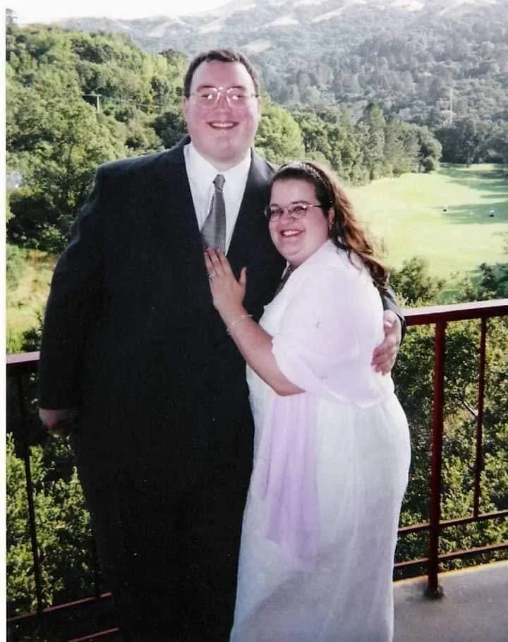 A smiling couple standing outdoors on a balcony with a scenic green landscape and hills in the background. The man is wearing a black suit with a tie, and the woman is in a white dress with glasses, embracing each other.