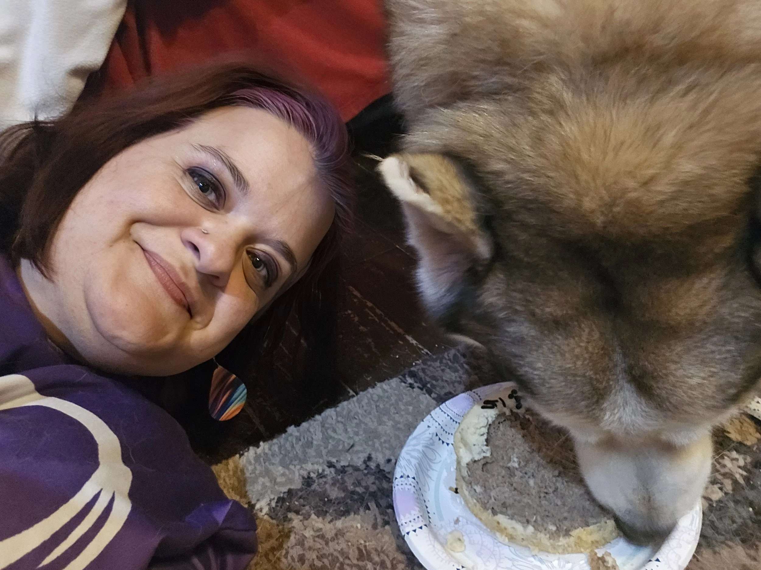 A woman with short brown hair and a nose piercing is lying on the ground next to a large brown and black dog. They are both laying on a textured surface, possibly an outdoor patio or concrete. The woman is smiling and looking at the camera, while the dog is resting its head near her. There is a paper plate with a partially eaten piece of food on it, placed in front of the dog.