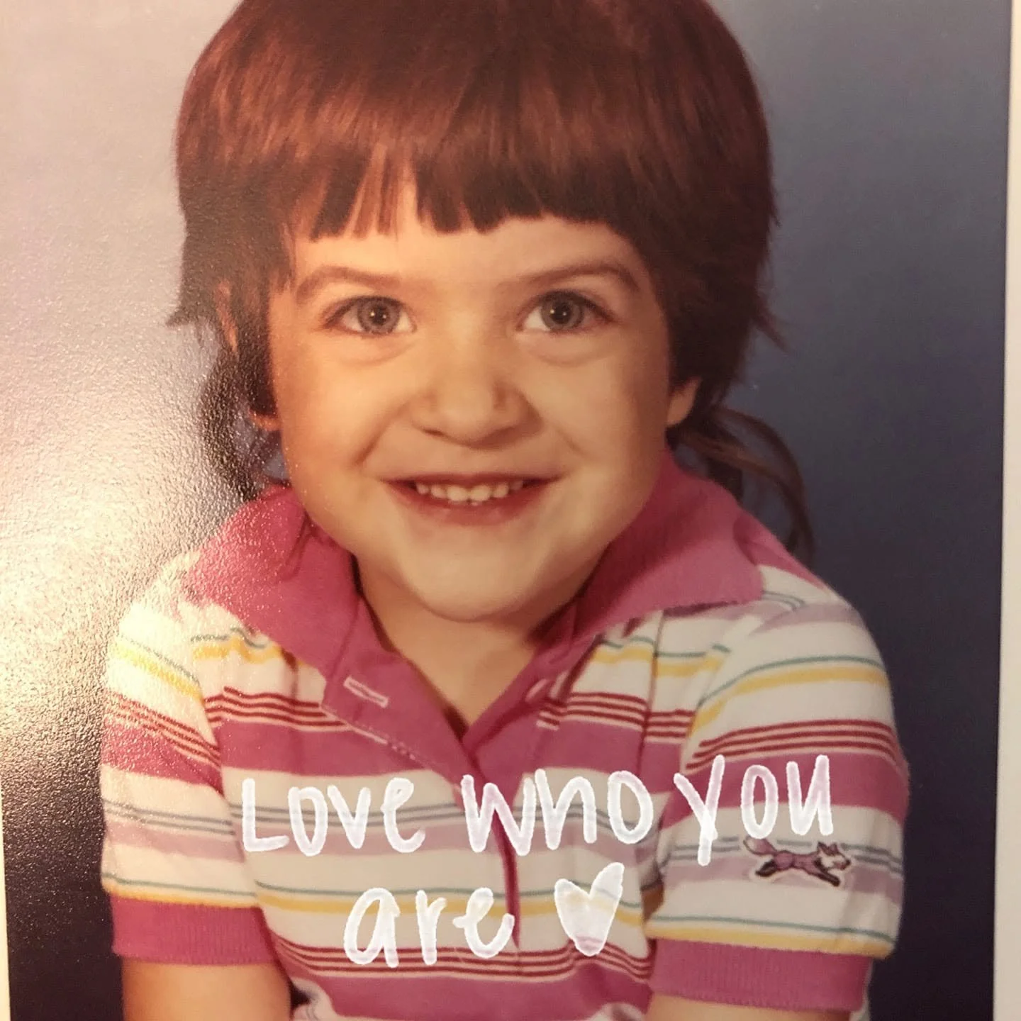Smiling young girl with short brown hair, wearing a pink and white striped polo shirt, and the text 'Love who you are' with a heart graphic.