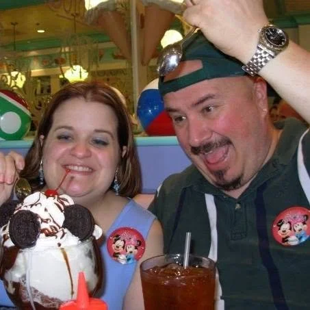 A smiling woman and man celebrating, sitting at a table with desserts and drinks, in a colorful restaurant or amusement park setting.