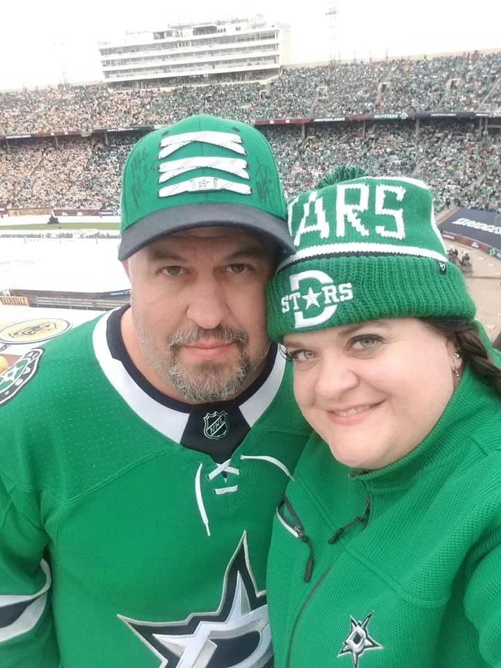 A man and woman at a sports stadium in Dallas, Texas, wearing Dallas Stars hockey team gear, smiling for a selfie with a large crowd in the background.