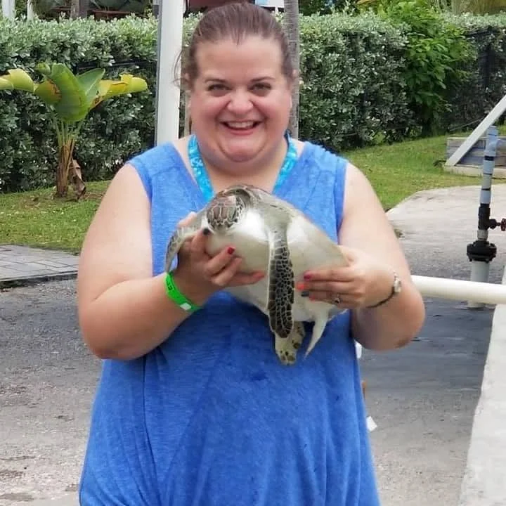 A smiling woman in a blue sleeveless shirt holding a small sea turtle outdoors.