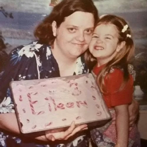 A woman and a young girl smiling and hugging, holding a pink birthday cake with the name 'Lila' written on it, in a cozy indoor setting.